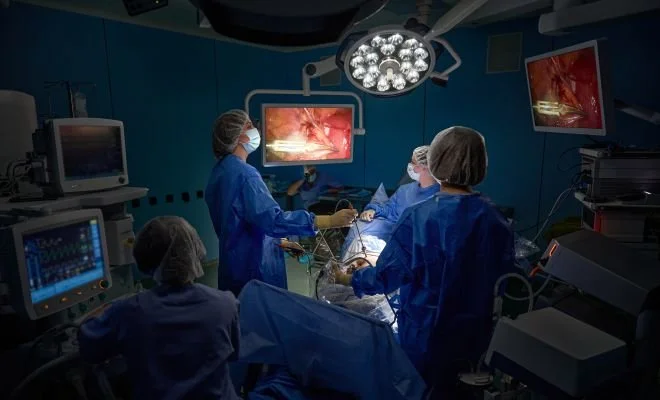 Surgeons and medical staff performing surgery in an operating room with monitors displaying surgical images.