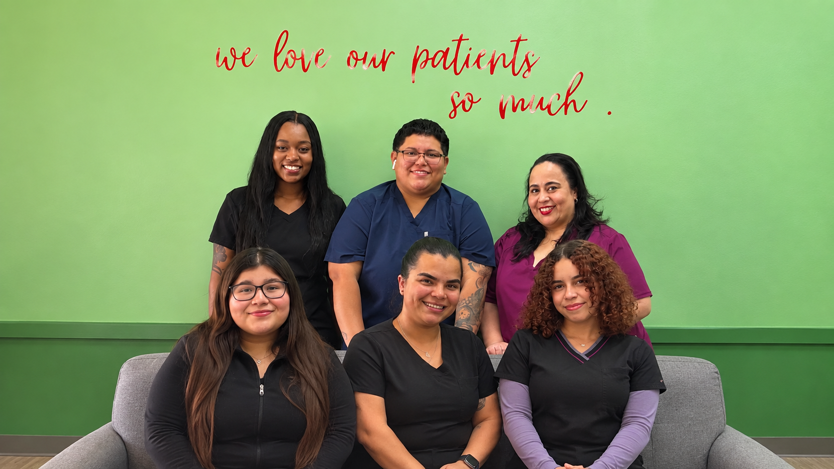 Group of six healthcare workers posing in front of a green wall with red cursive text reading 'we love our patients so much.'