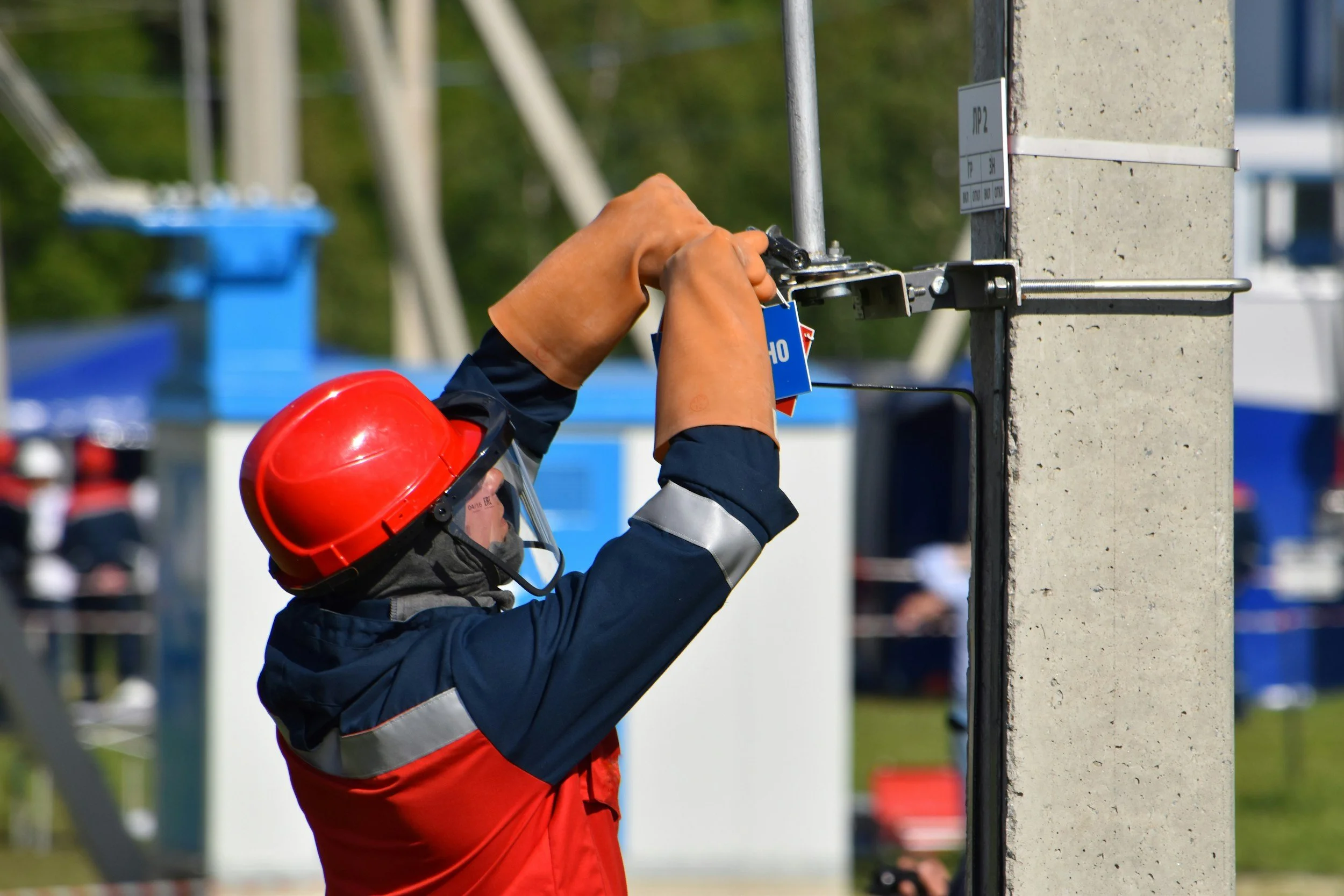 Worker wearing a red helmet and safety gear repairing or installing equipment on a concrete column at an outdoor construction site.