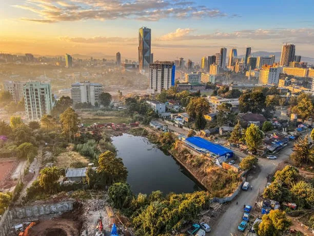 City skyline during sunset with high-rise buildings, a small lake, green trees, and streets with cars in foreground.
