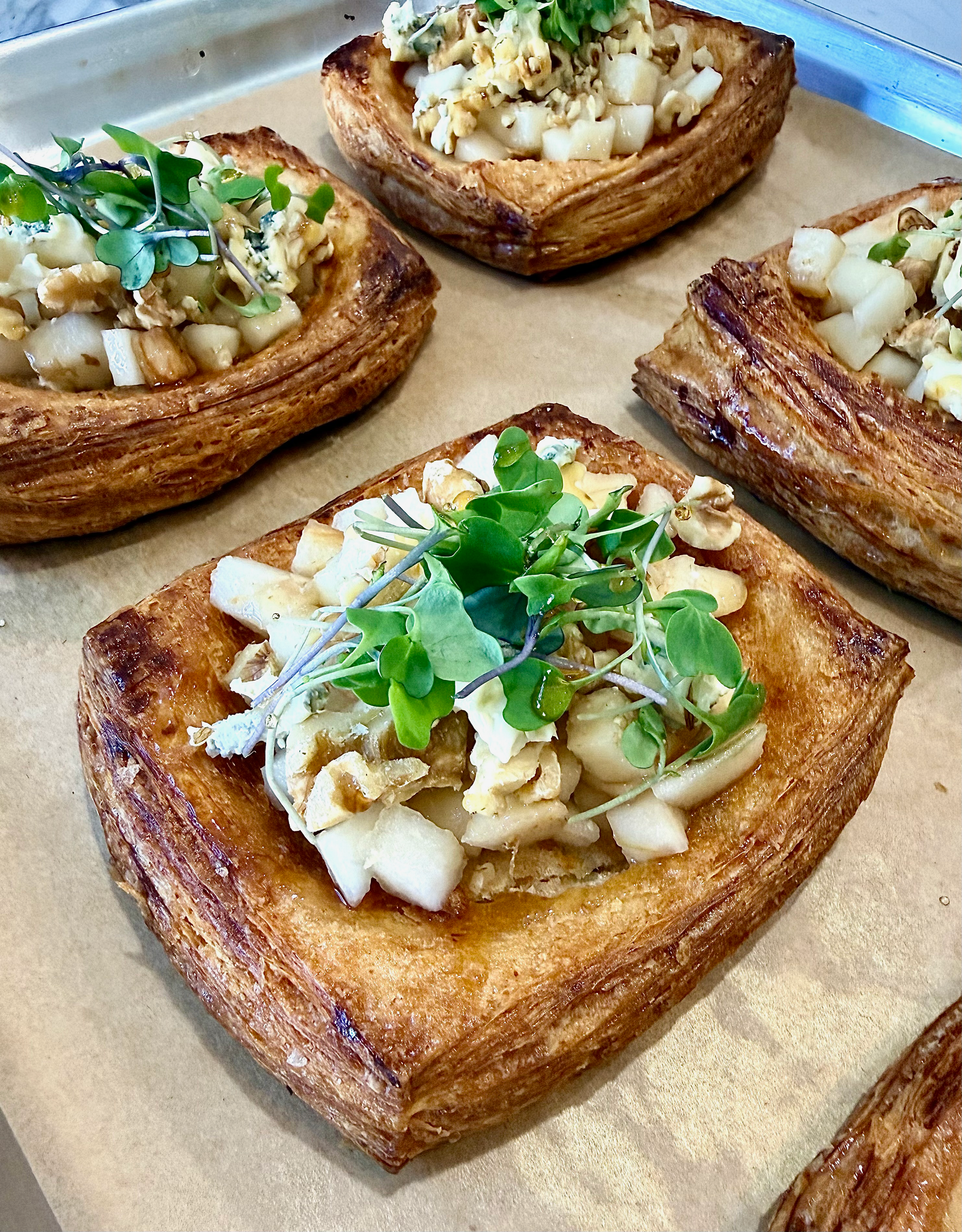 Close-up of puff pastry squares topped with chopped vegetables, microgreens, and cheese on a parchment-lined tray.