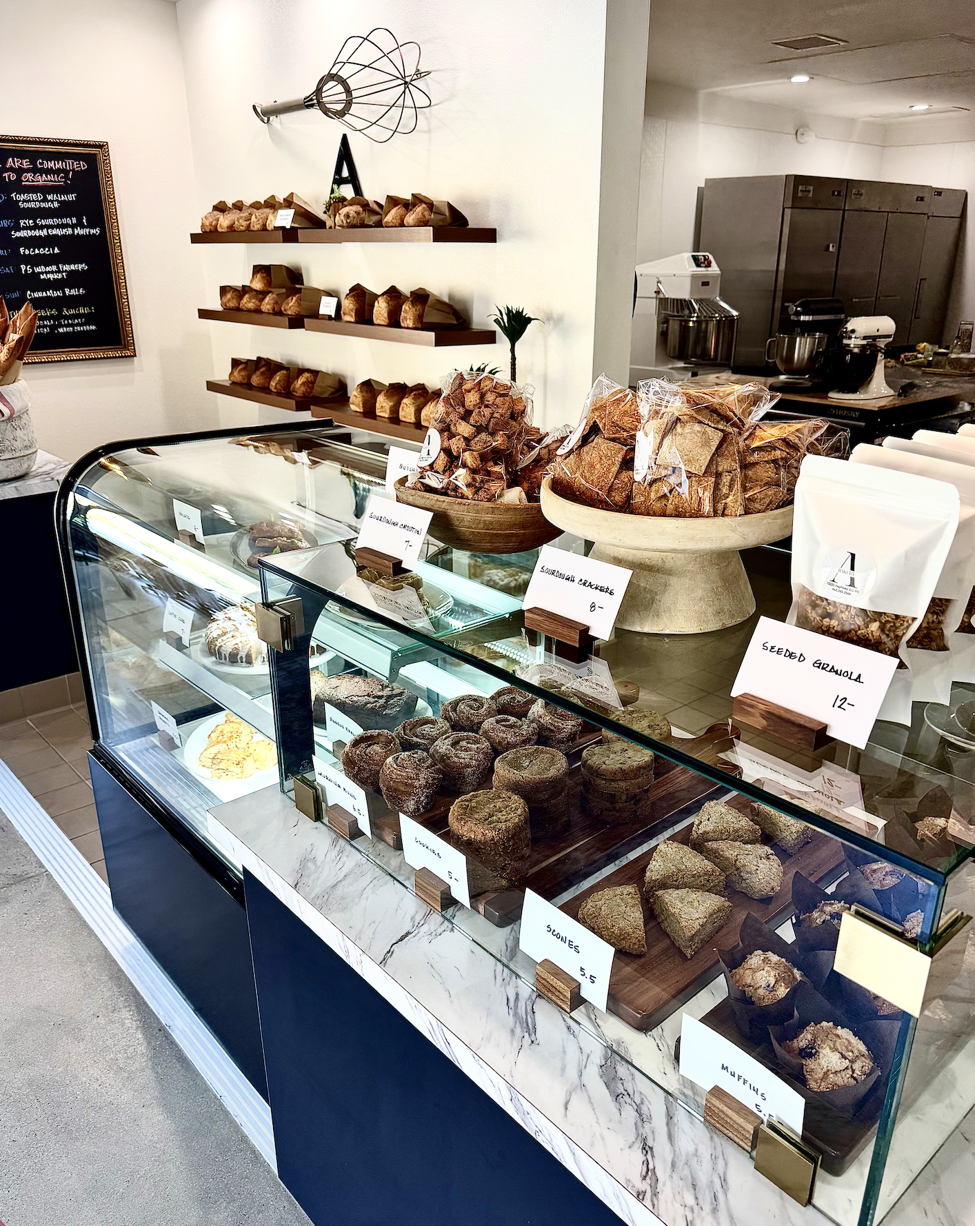 Display case filled with various baked goods, including scones, muffins, and granola. Behind the counter, shelves hold loaves of bread and baked treats. A chalkboard menu lists bakery items, with some packaging and signs showing prices.