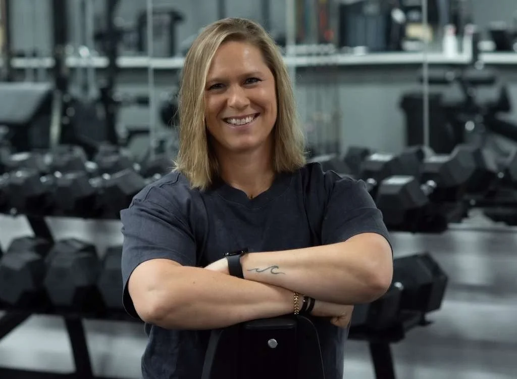 A smiling woman with shoulder-length blonde hair standing in a gym with dumbbells in the background.
