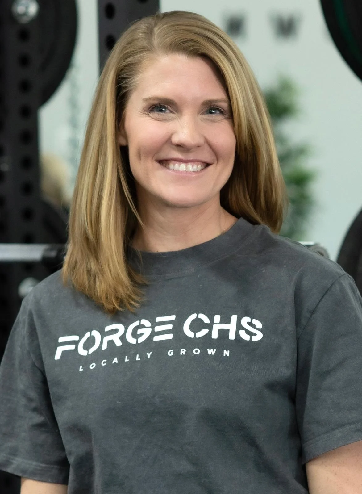 A smiling woman with shoulder-length blonde hair, wearing a dark gray T-shirt with the words 'FORGE CHS LOCALLY GROWN' printed on it, standing in what appears to be a gym or fitness center.