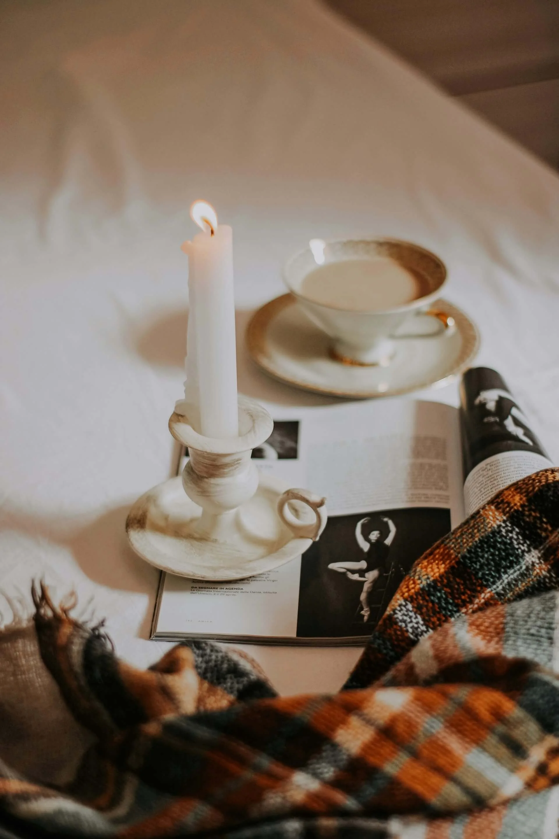 A lit white candle on a decorative candle holder, a teacup with a saucer, a magazine or book with black-and-white photos and text, a partially visible animal (possibly a dog), an orange and black plaid fabric, and a black bottle.