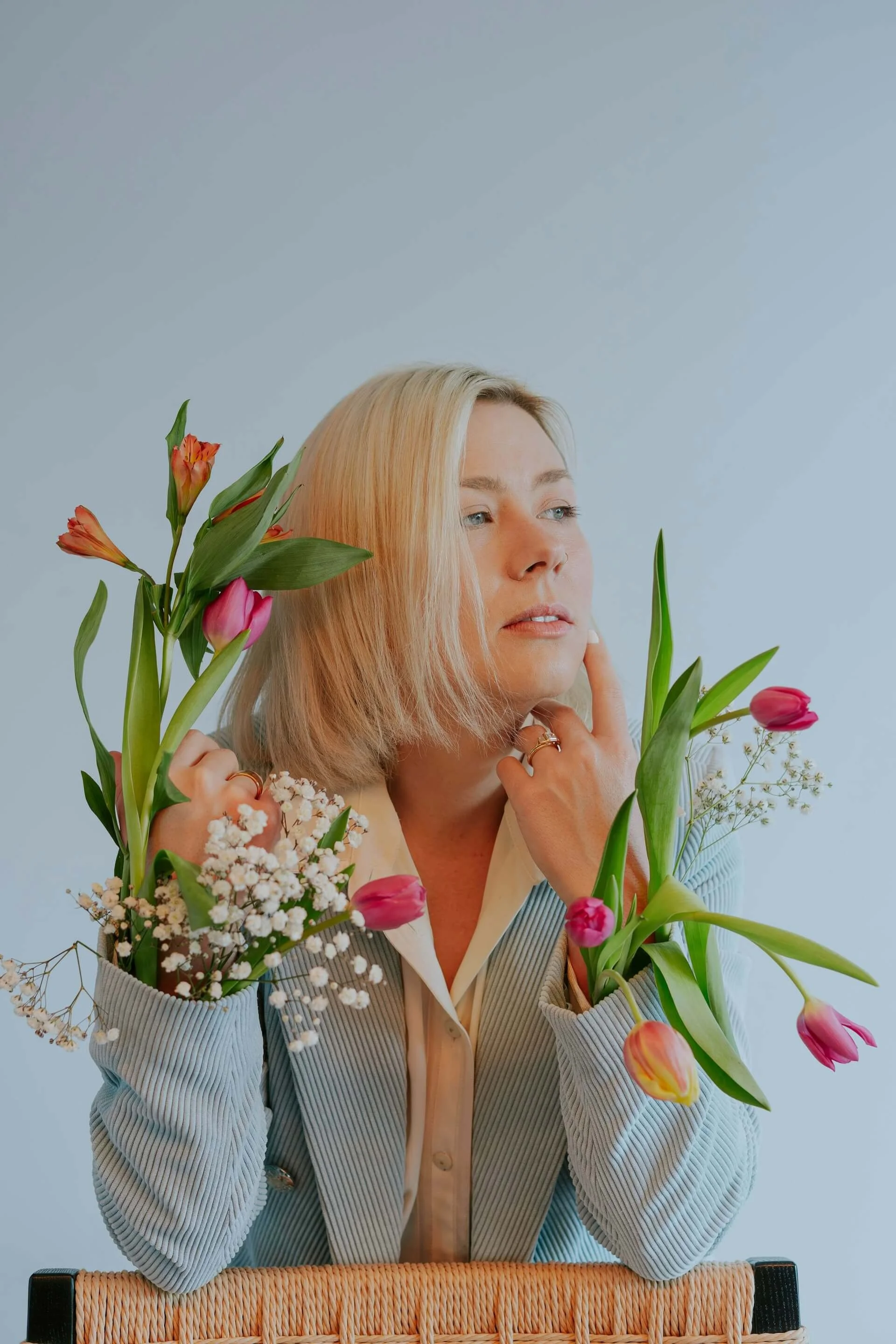 A woman with blonde hair, wearing a striped blazer and beige top, surrounded by various colorful flowers including tulips and baby's breath, sitting in front of a plain light blue background.