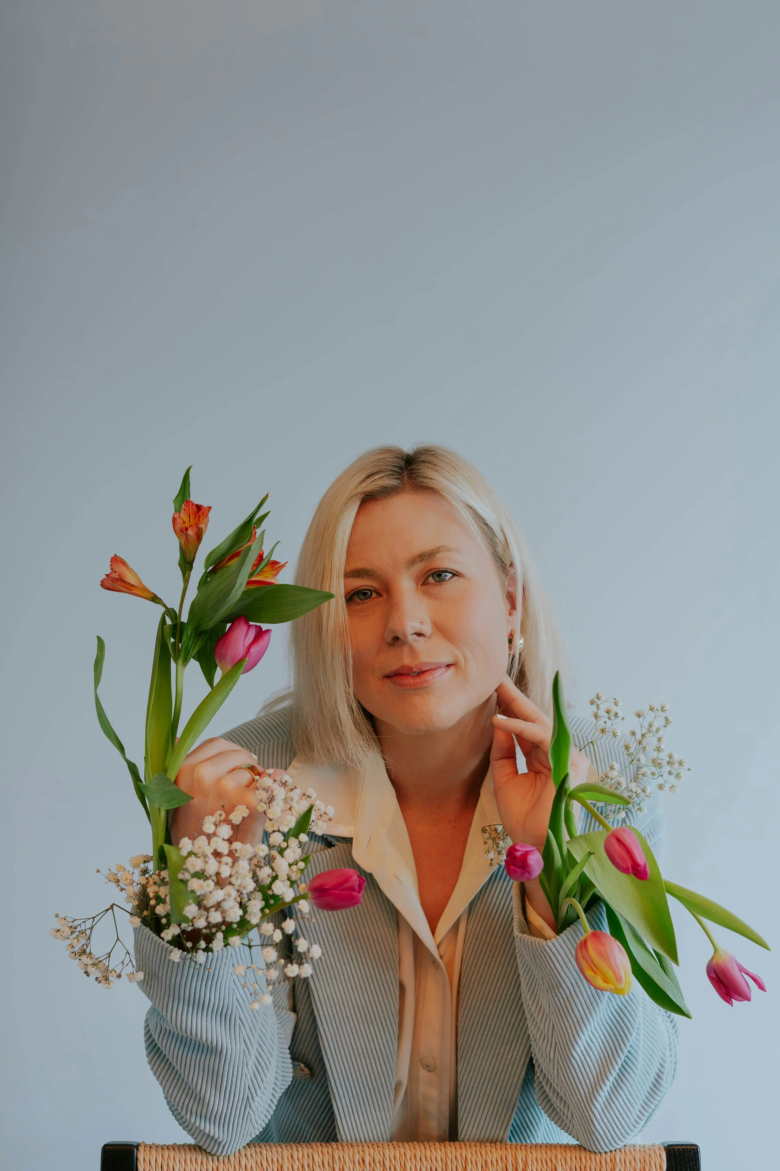 A woman with blonde hair sitting at a table, holding a bouquet of various flowers, including pink tulips and white daisies, with a plain light blue background.