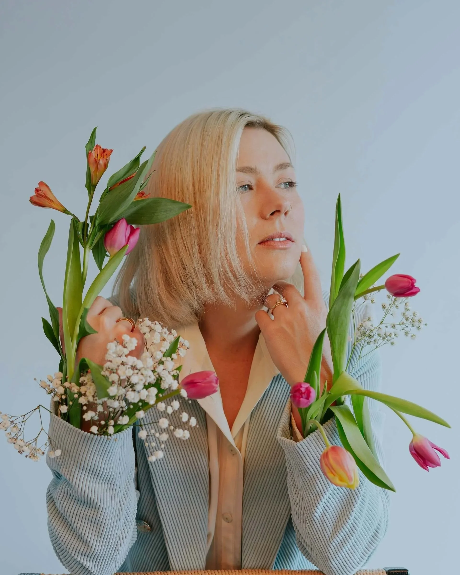 A woman with blonde hair holding colorful tulips and white baby's breath flowers, wearing a beige and blue striped cardigan and a white button-up shirt, sitting at a table against a plain light blue background.