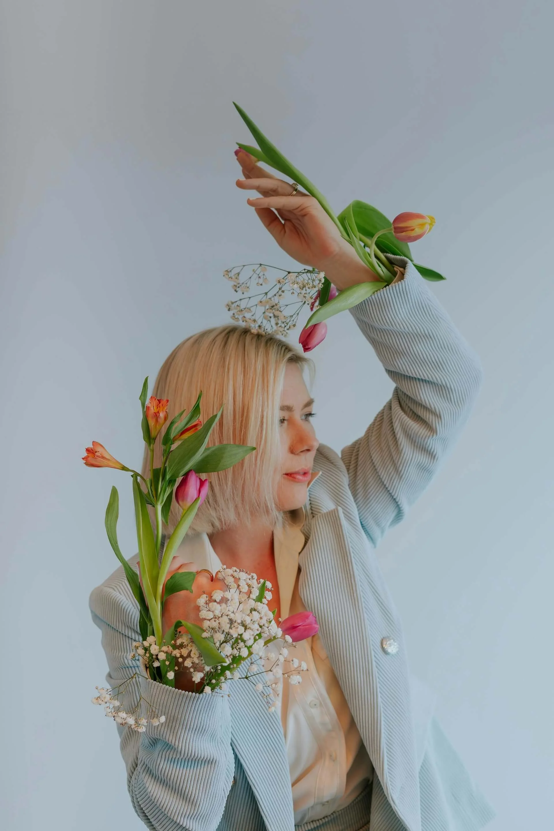 Woman with blonde hair holding a bouquet of colorful flowers, including tulips and baby's breath, against a plain background.