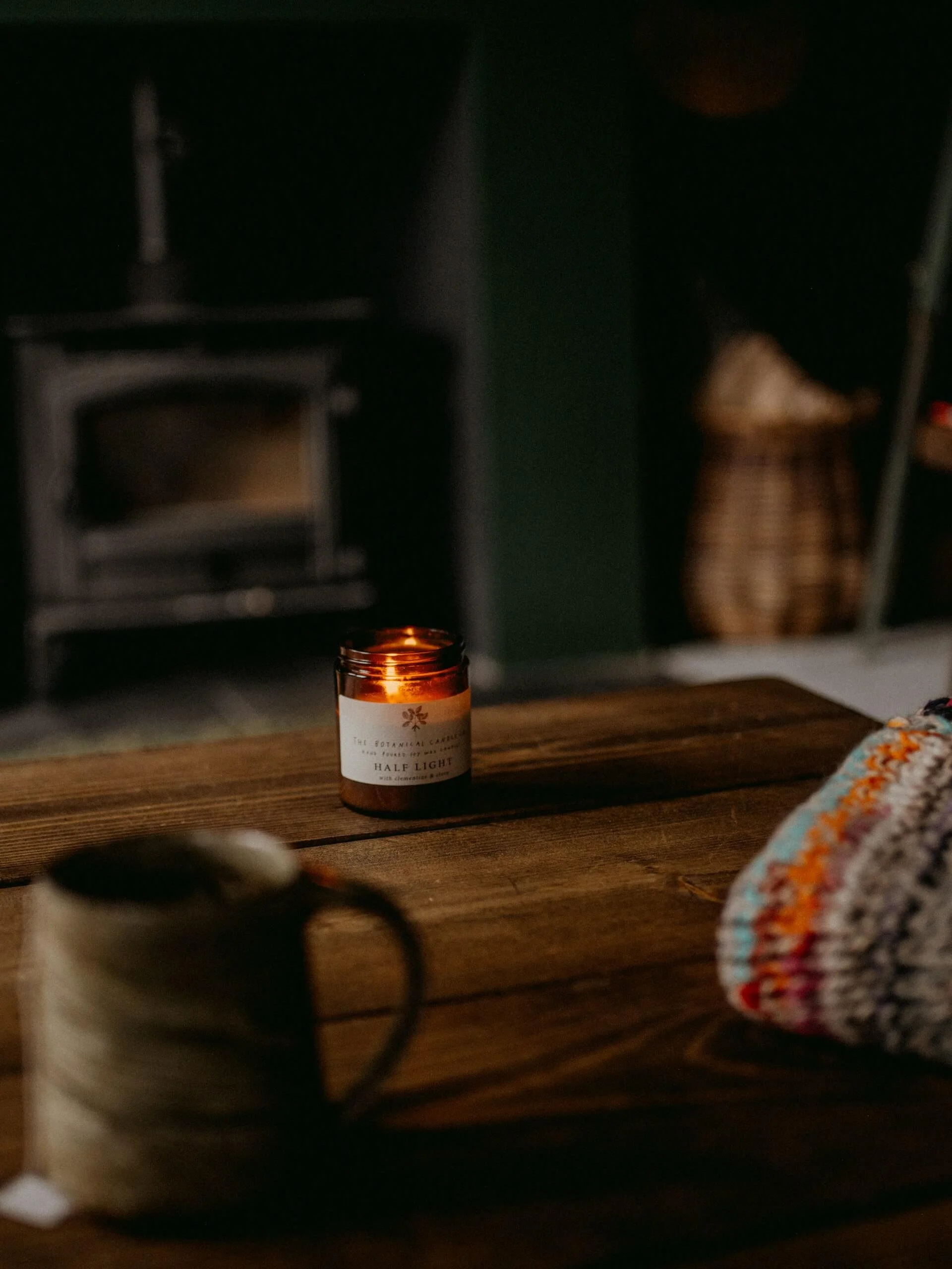 A cozy room with a wooden table, a lit candle, and a colorful cloth or blanket on the right side. The background includes a dark wall, a fireplace, and a person sitting on a chair.