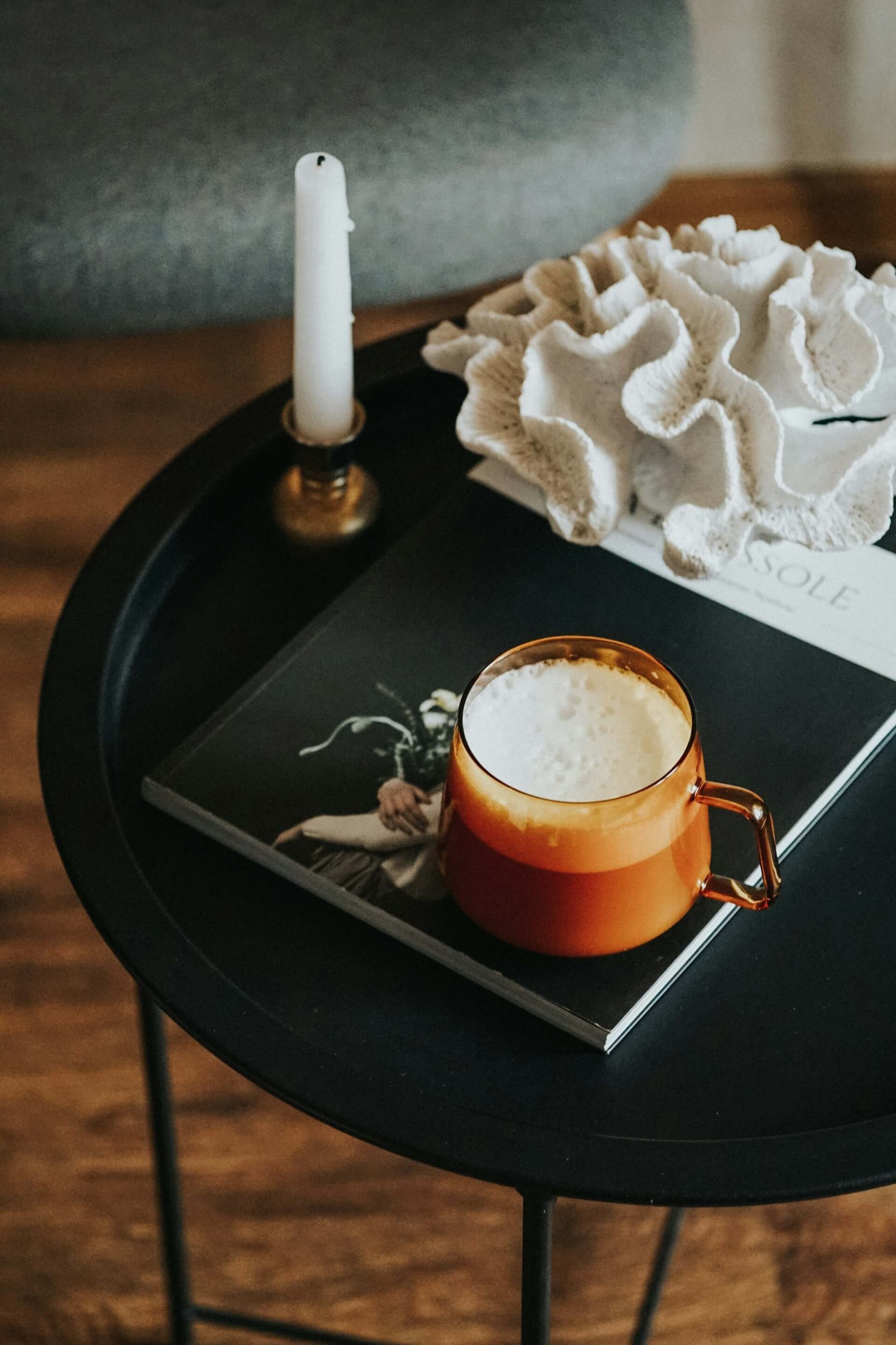 Black round table with a candle, a large white decorative object, a book, and an amber-colored mug filled with a frothy beverage.