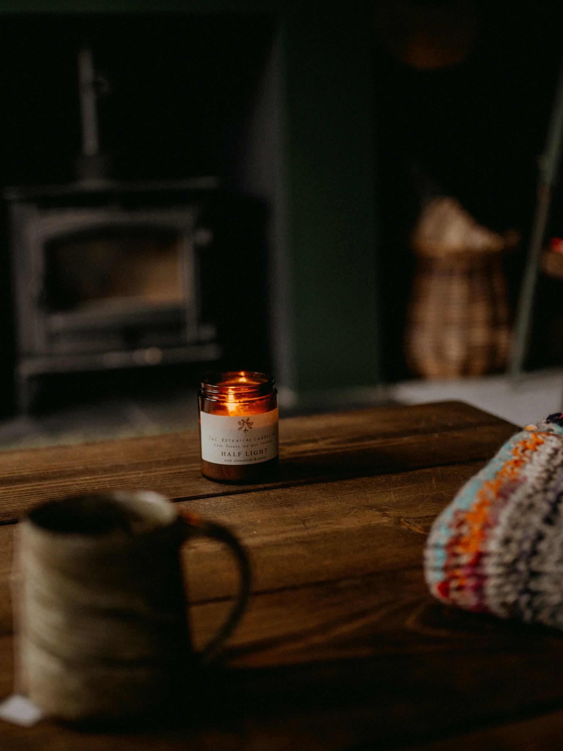 A lit candle on a wooden table with a label reading 'Half Light' in a dimly lit room. In the foreground, part of a mug and a colorful cloth are visible.