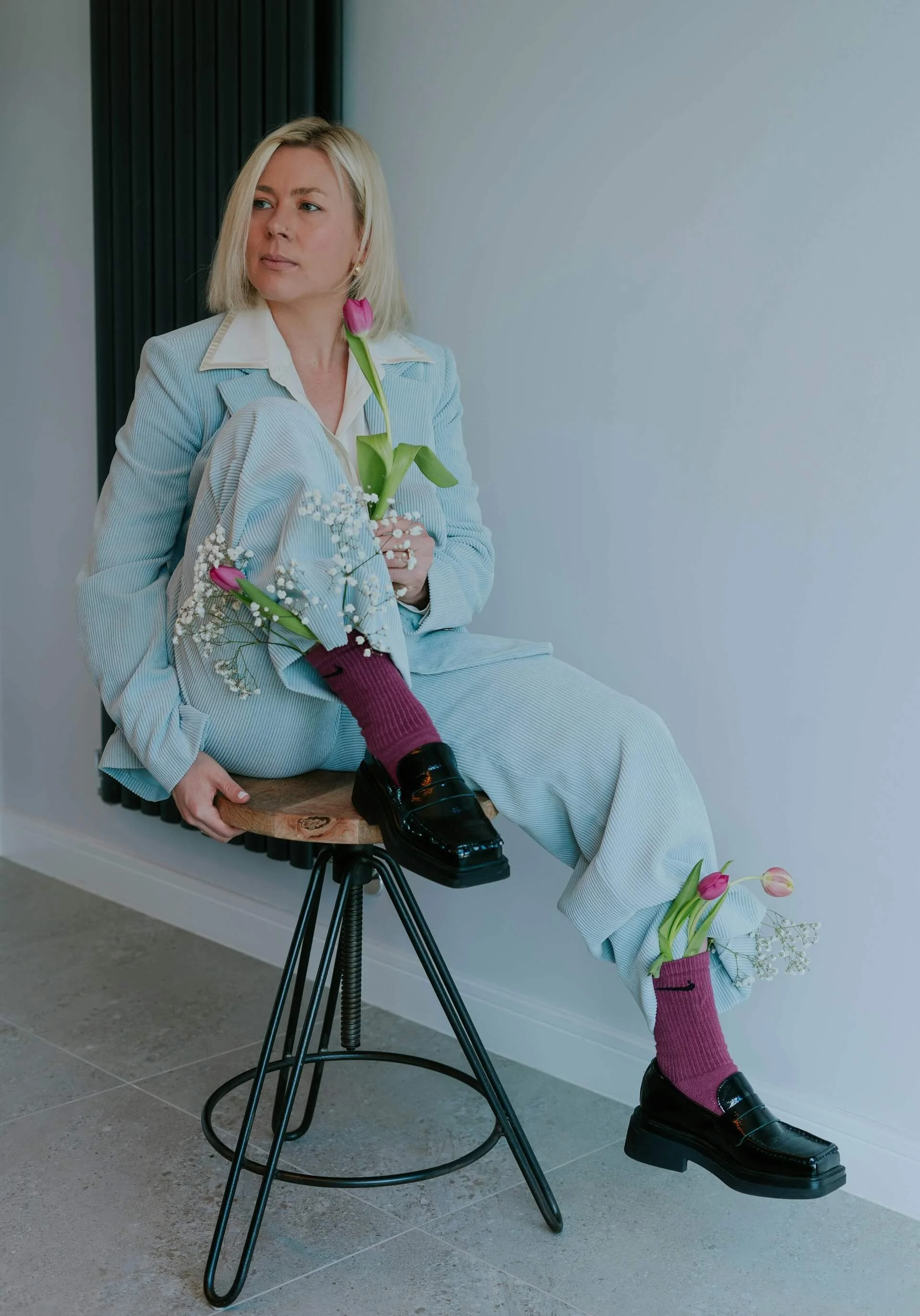 A woman in a light blue pinstripe suit and white shirt sitting on a stool, holding flowers, with one ankle resting on her knee, against a plain wall.