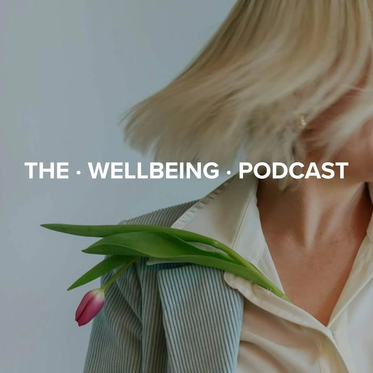 Close-up of a woman with blonde hair, wearing a light-colored blazer and white shirt, with a pink tulip and green leaves resting on her shoulder. Text overlay reads 'The Wellbeing Podcast'.