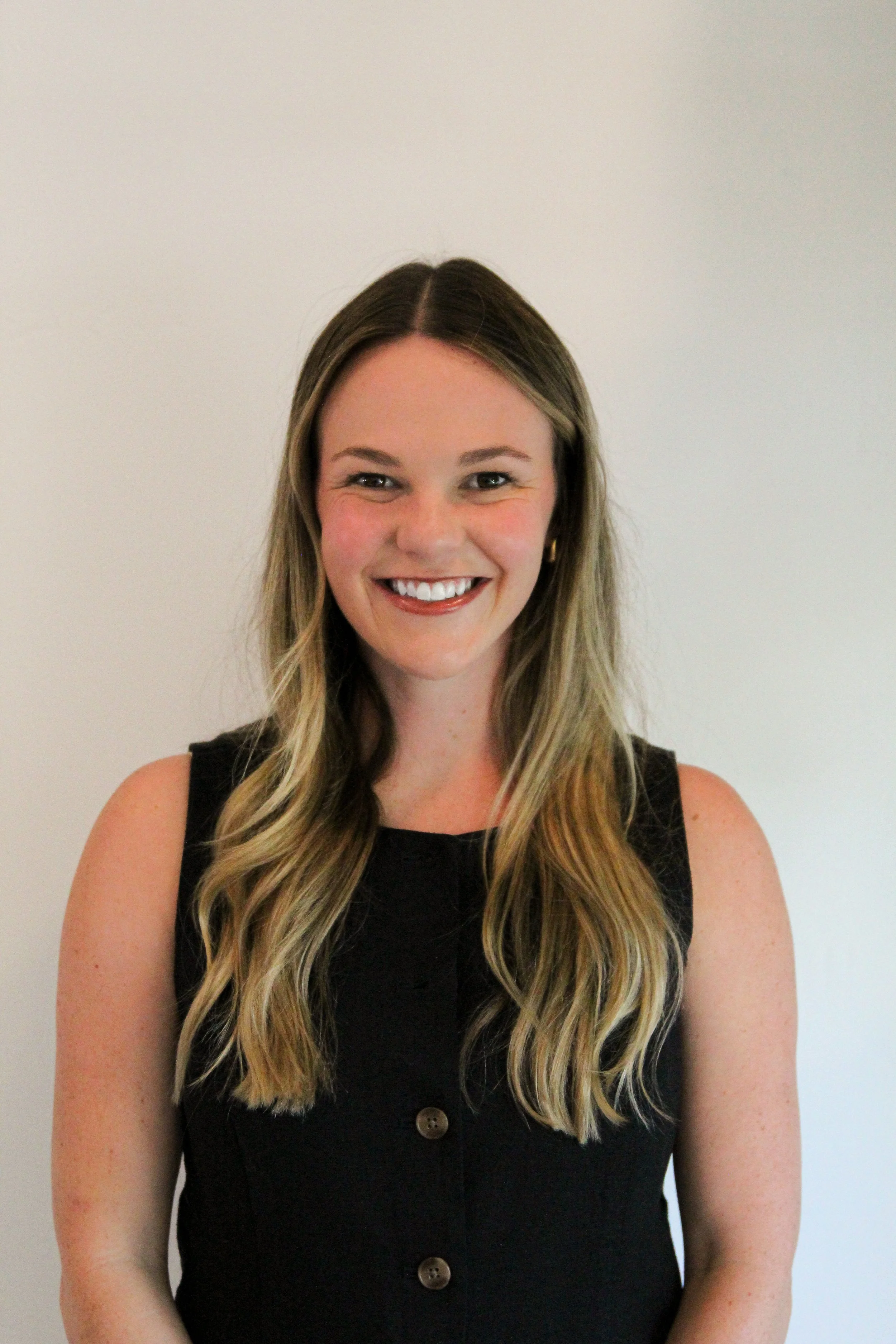 A smiling woman with long, wavy blonde hair wearing a sleeveless black top, standing against a plain white wall.