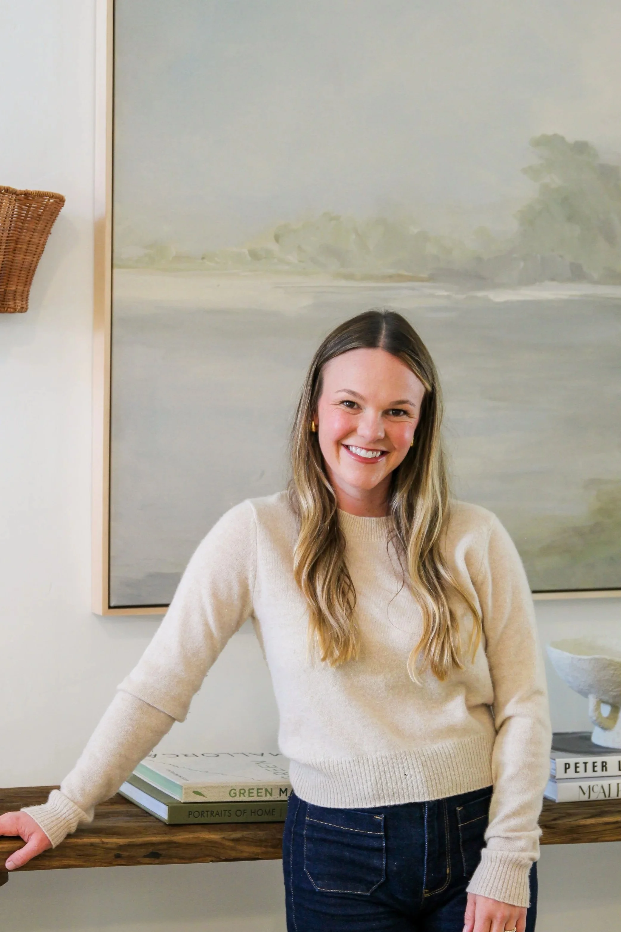 A woman with long wavy blonde hair, wearing a cream-colored sweater and dark blue jeans, is smiling at the camera. She is standing in front of a wooden table with books on it, behind her is a wall with a large landscape painting, and a basket is mounted on the wall to her left.