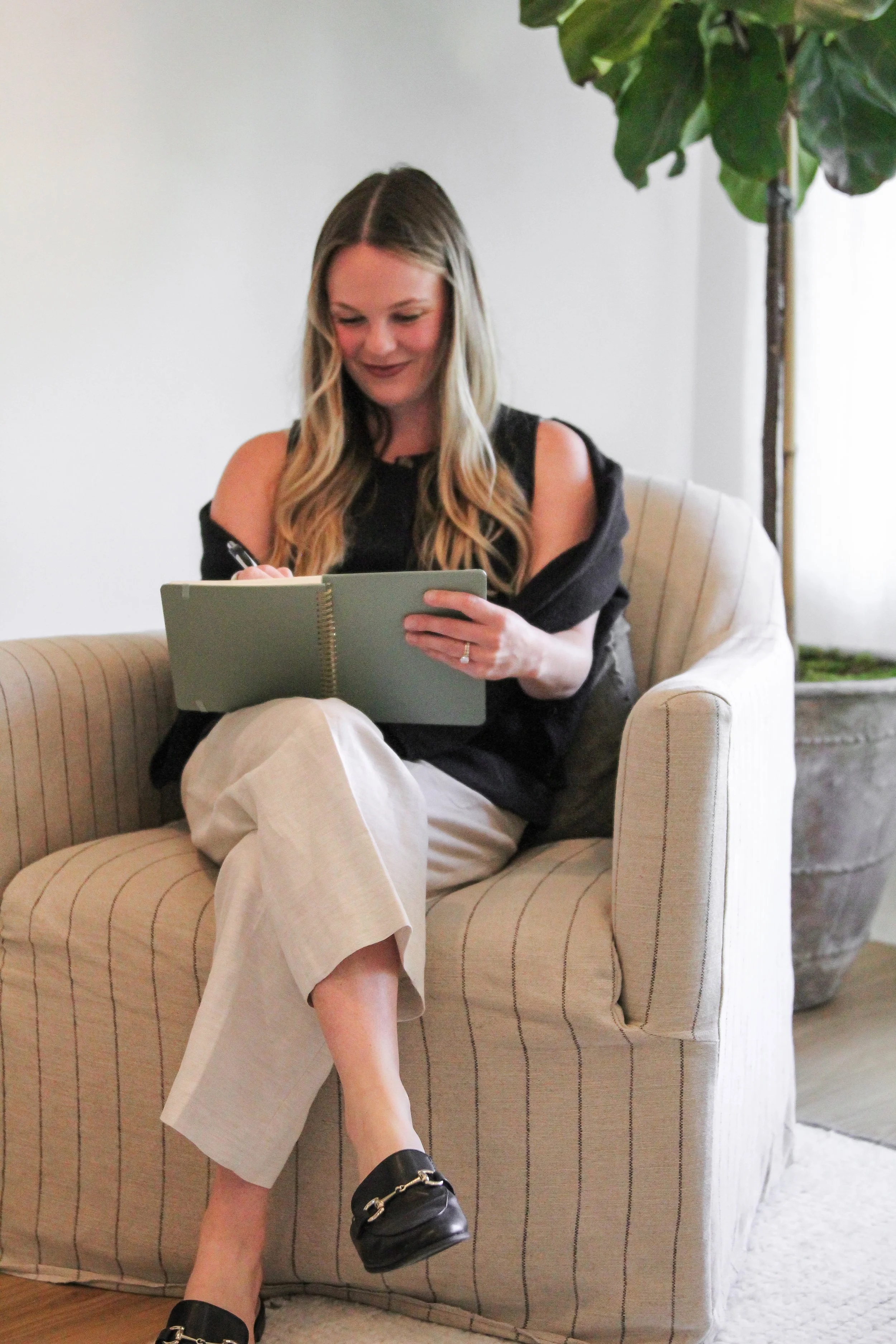A woman with long wavy blonde hair sitting on a beige striped armchair, writing in a green notebook. She wears a black top with torn shoulders, beige pants, and black loafers with gold chain detail, in a modern living space with a large green leafy plant in the background.