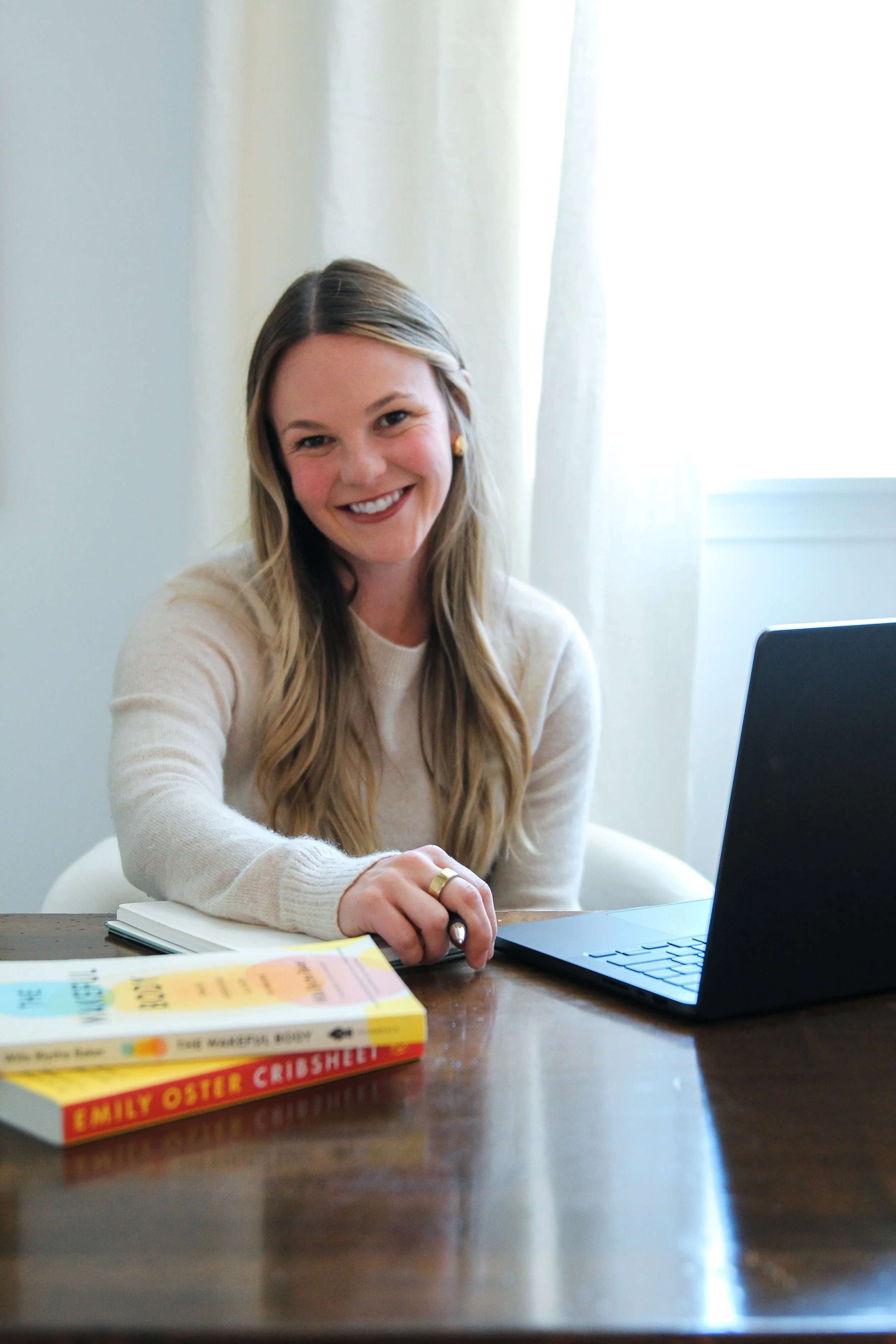 A woman with long blonde hair, wearing a light-colored sweater, smiling while sitting at a wooden table with a laptop and a book titled 'Family Oster Crisbhet' in front of her. There are light-colored curtains and a window in the background.