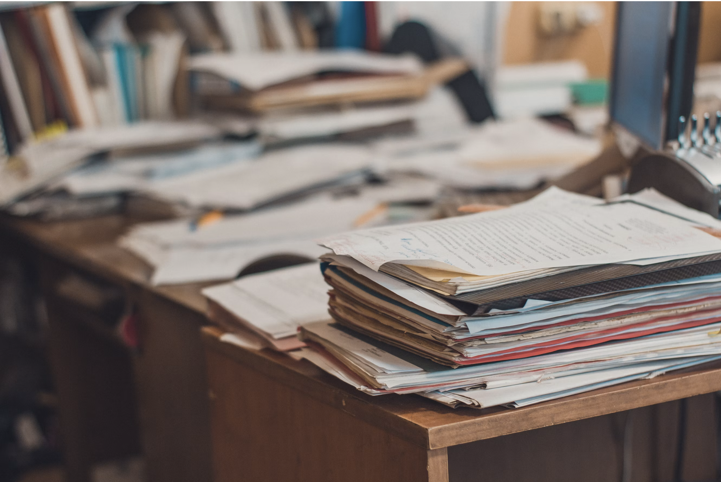A cluttered desk filled with stacks of paper, files, and documents, with a bookshelf in the background.