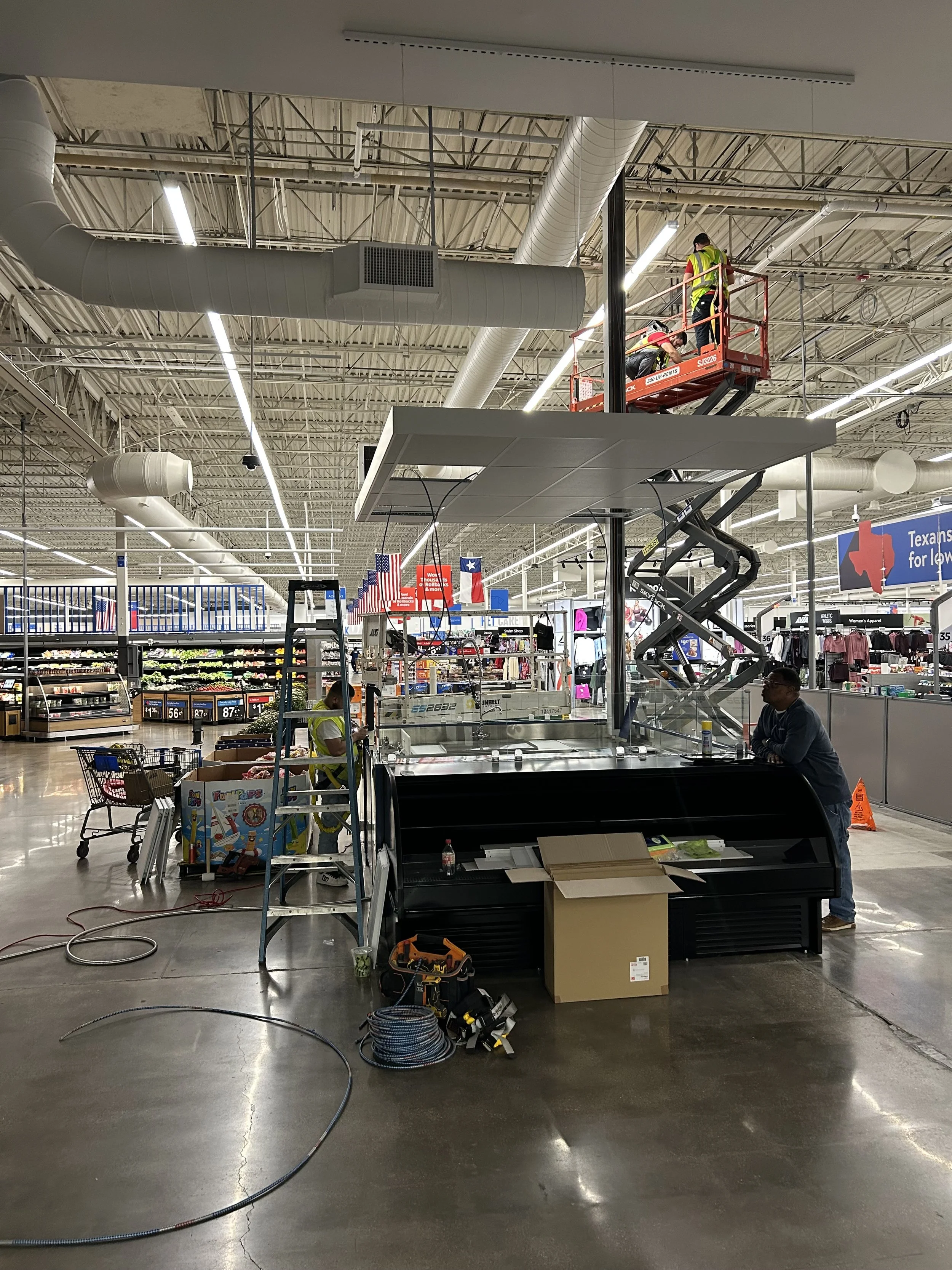 Inside a store, two workers in high-visibility vests are on a scissor lift working on the ceiling, which has exposed ductwork and lighting fixtures. The store appears to be under renovation or maintenance.