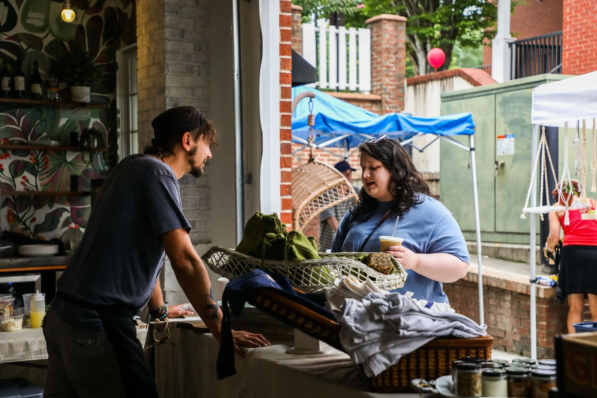 A man and a woman are at an outdoor market stall, engaging in a conversation. The man is leaning on the counter, and the woman is holding a drink and looking at the man. The stall has various items, including a basket of clothes and other goods. There are tents and other market stalls in the background, along with brick walls and trees.