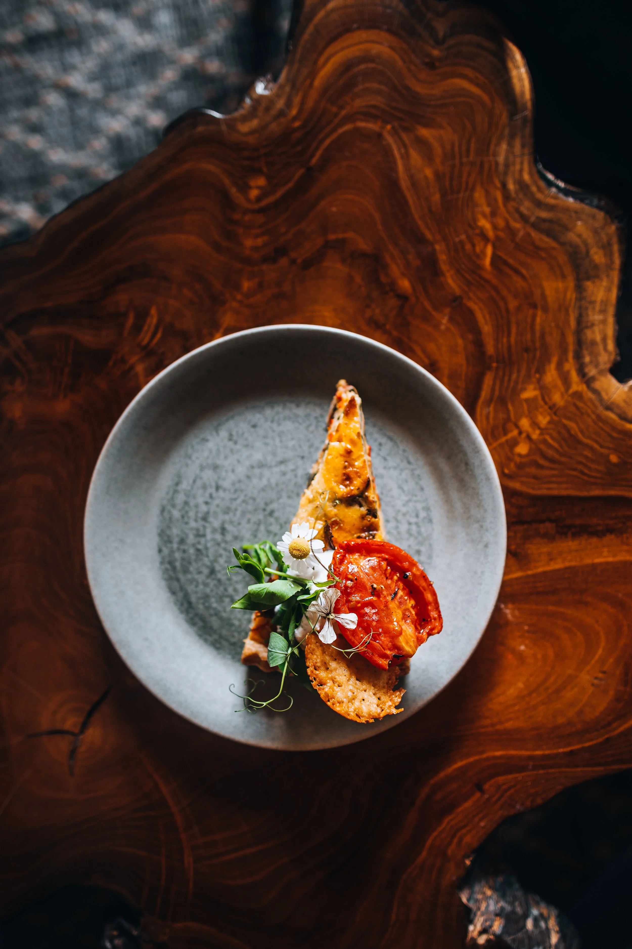 A slice of pizza on a gray plate with green herbs and small white flowers, resting on a wooden table.