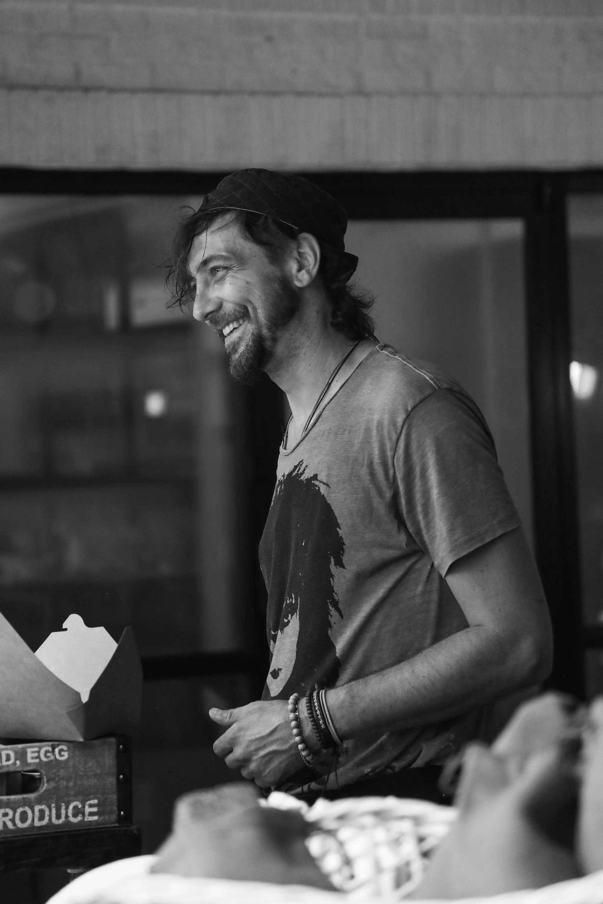 A man with long hair, a beard, and a smiling expression is wearing a cap, a t-shirt with a graphic, and multiple bracelets and wristbands. He is standing behind a counter, possibly serving or preparing food, with a box of eggs labeled 'PRODUCE' on it. His face is turned slightly to the side. The background appears to be an indoor setting with wooden or brick walls.
