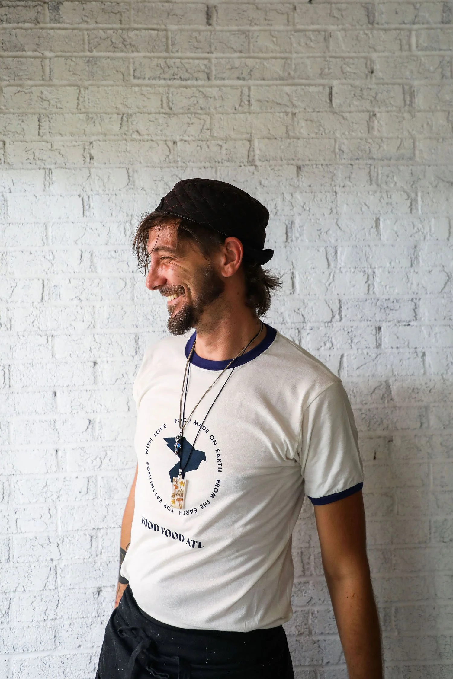 A man with long hair, a beard, and a mustache, smiling joyfully, wearing a black cap, a white T-shirt with a circular design and text, and black pants, standing against a white brick wall.