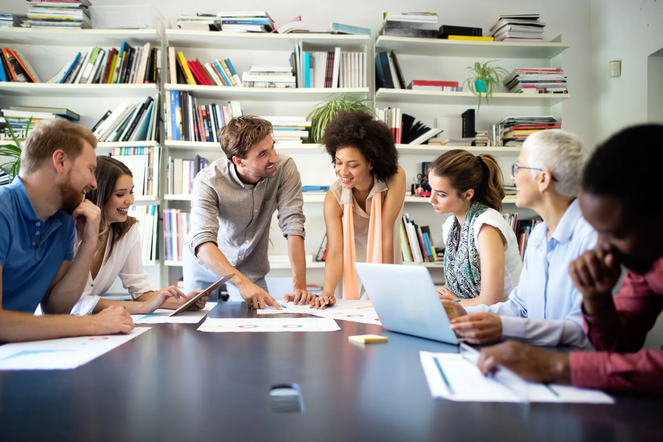 Group of diverse people around a table in a meeting or collaborative work session, with bookshelves in the background.
