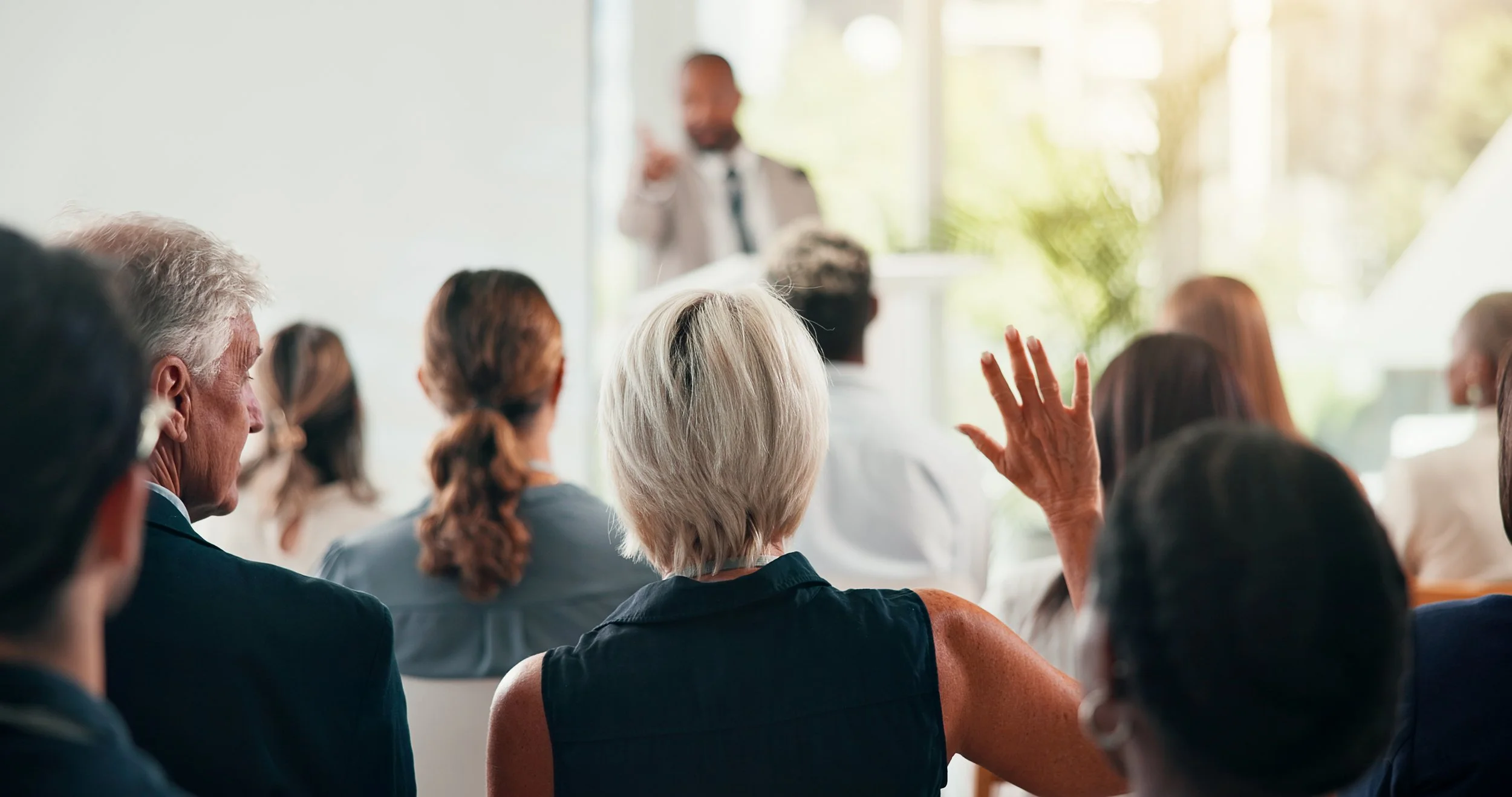 A group of diverse people attending a presentation or lecture, with one person raising their hand to ask a question, in a bright, modern room with large windows.