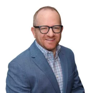A man with glasses, bald head, and a beard, smiling and wearing a blue blazer over a checkered shirt against a white background.