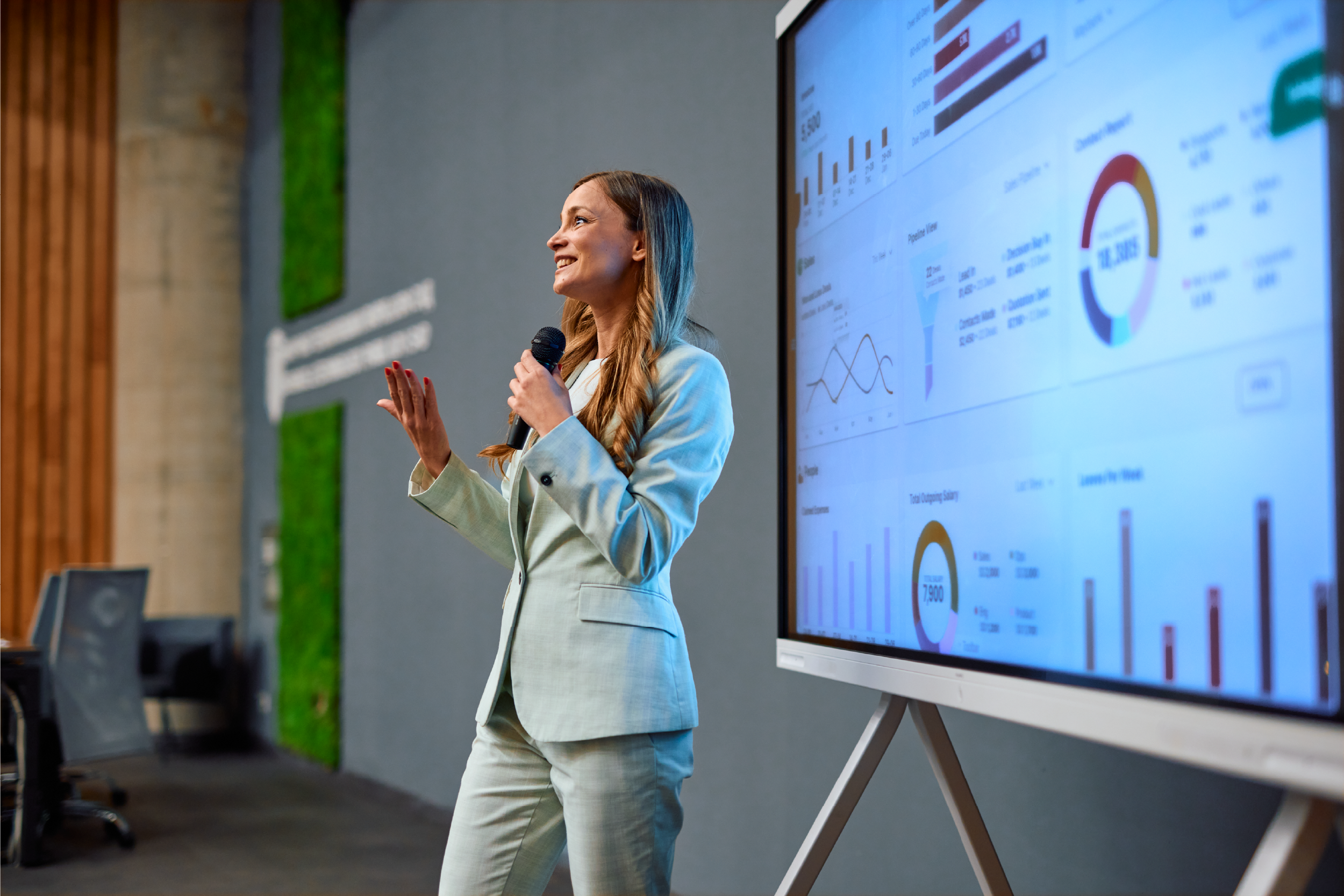 A woman in a light-colored suit giving a presentation with a microphone in front of a large screen displaying charts and graphs.