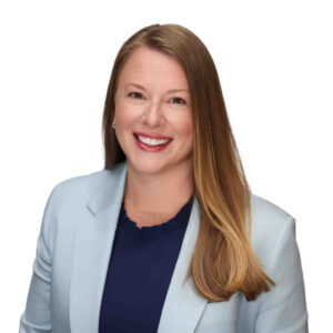 A smiling woman with long red hair wearing a light blue blazer and a navy top against a white background.