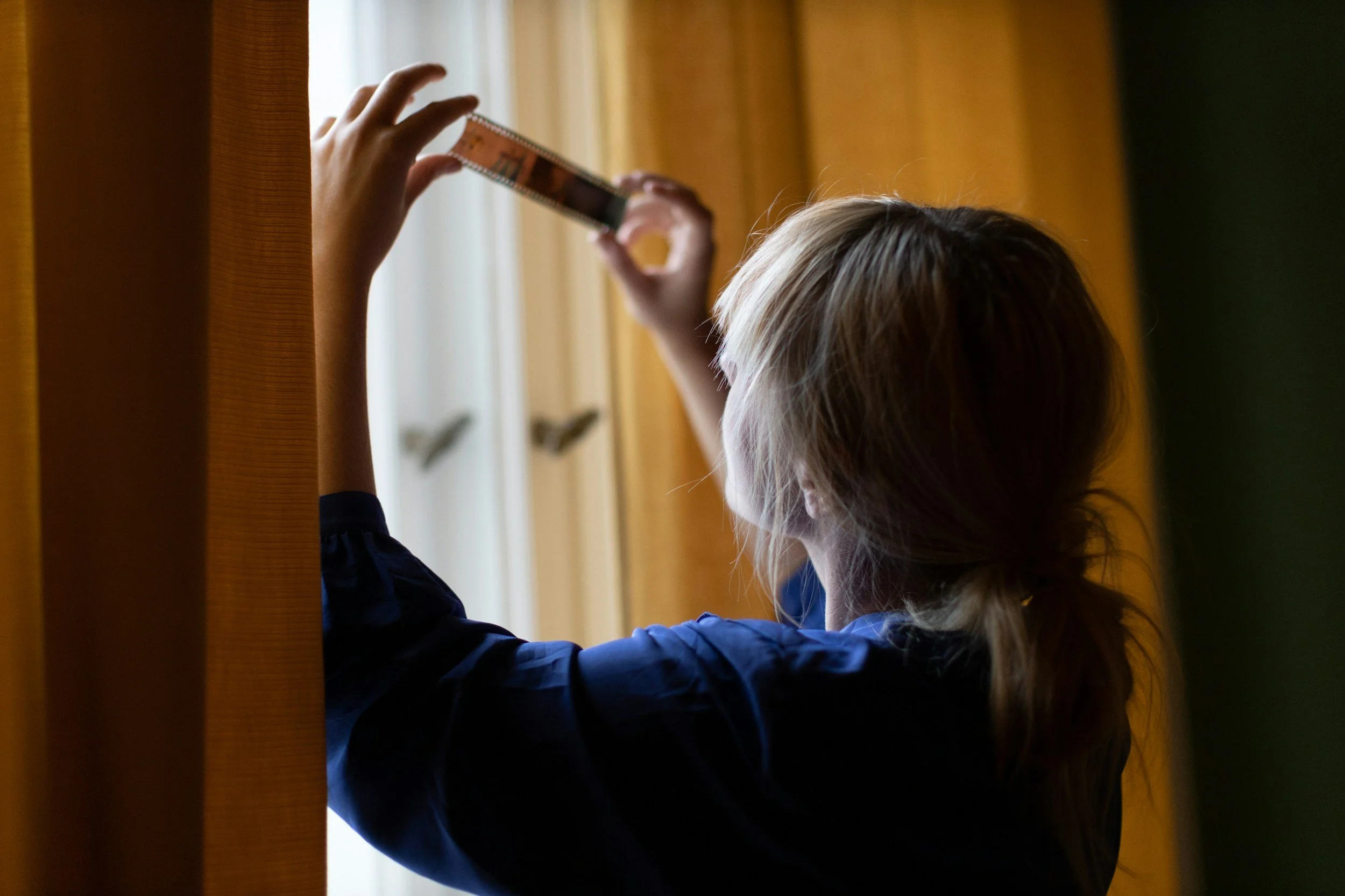 A child with blonde hair looking out a window while holding a strip of film up to the light.
