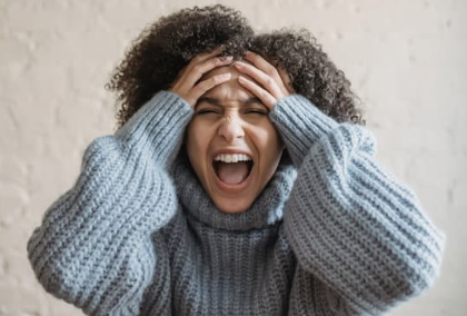 Woman with curly hair wearing a gray sweater, holding her head and screaming in frustration or distress.