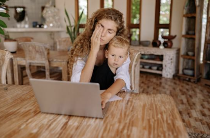 A woman and a young boy sitting at a wooden table looking at a laptop, with the woman appearing tired or stressed.