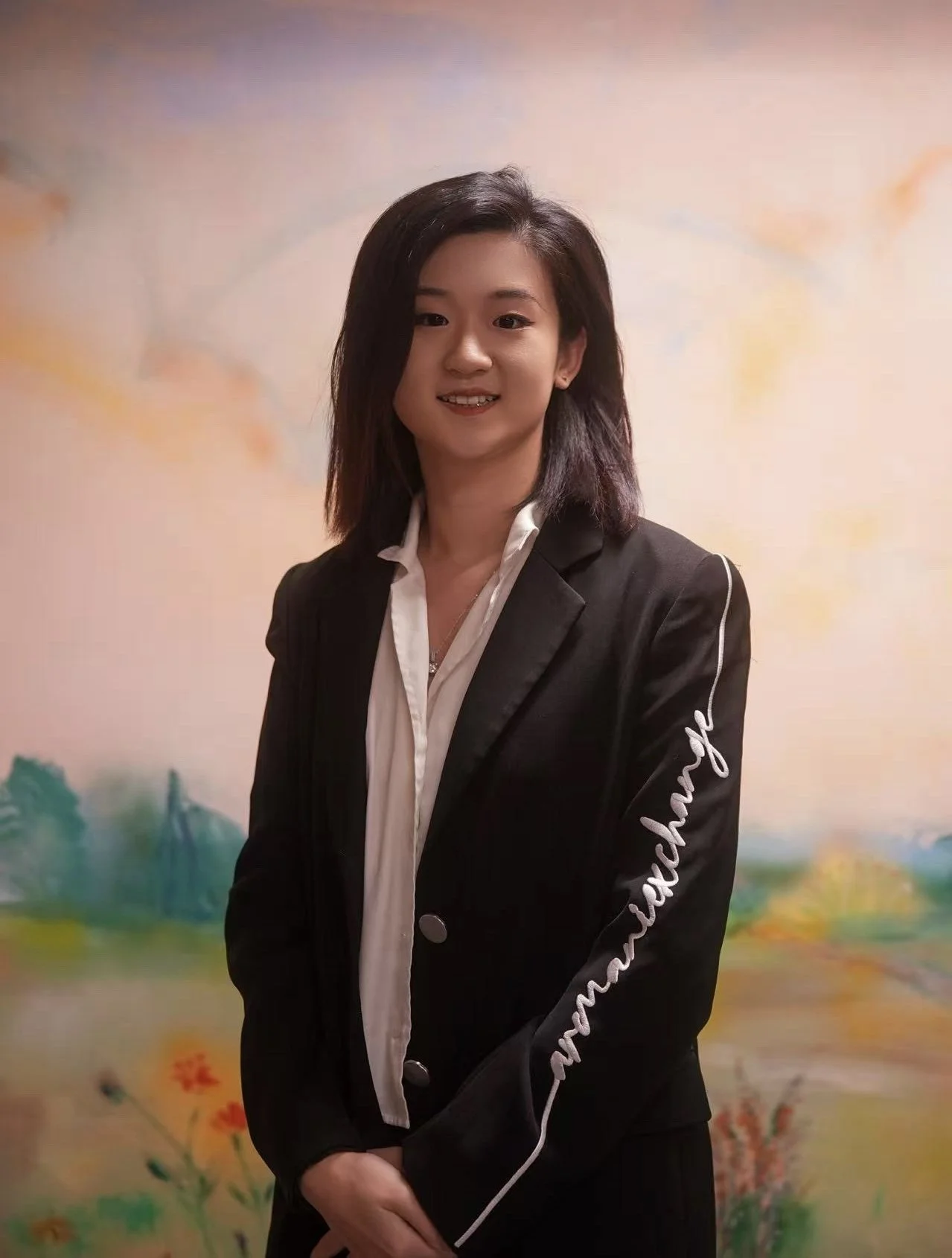 A young woman with shoulder-length black hair, wearing a black blazer with white cursive writing on the sleeve and a white collared shirt, standing in front of a blurred, colorful landscape background.