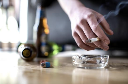 A person's hand with a cigarette above an ashtray on a wooden table, with a spilled bottle and small capsule in the background.