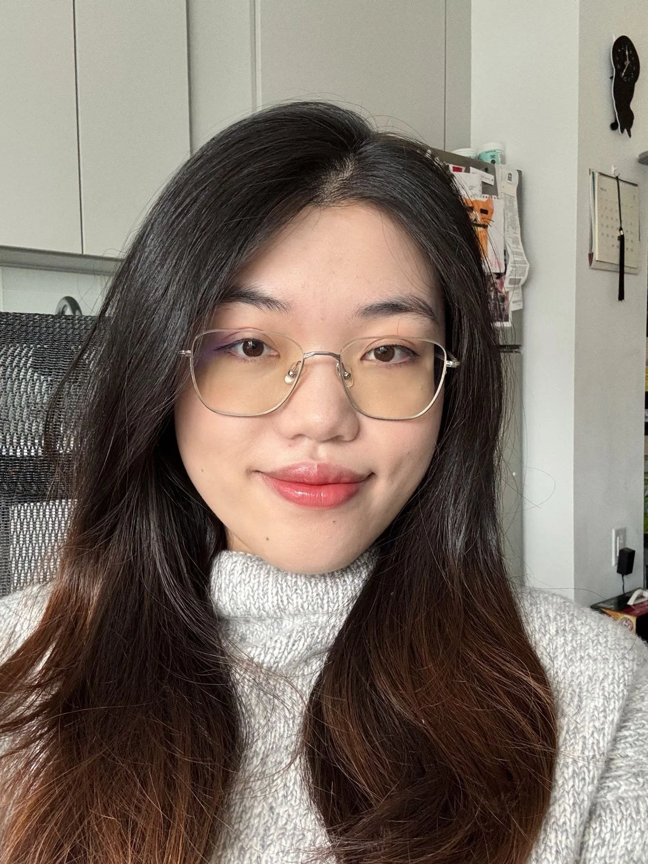 A young woman with long dark hair, glasses, and a white sweater smiling in an indoor setting.