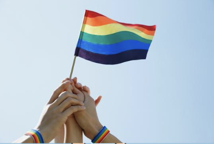 Two hands holding a rainbow pride flag against a light blue sky.