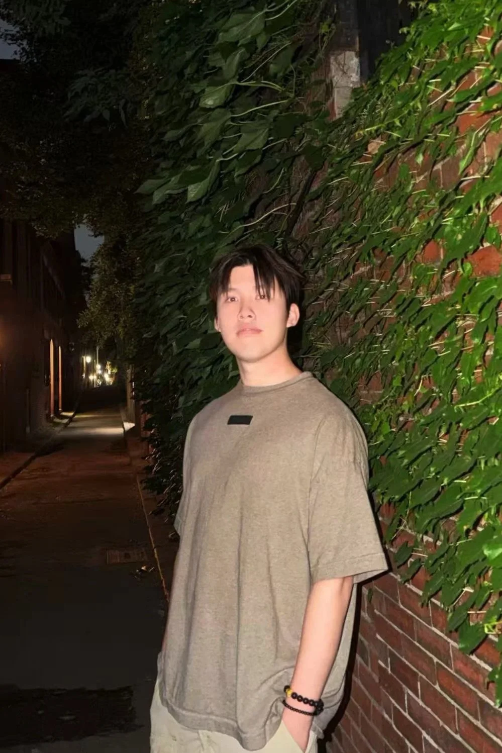 A young man standing near a brick wall with green foliage at night, wearing a beige oversized T-shirt and bracelets, looking at the camera.