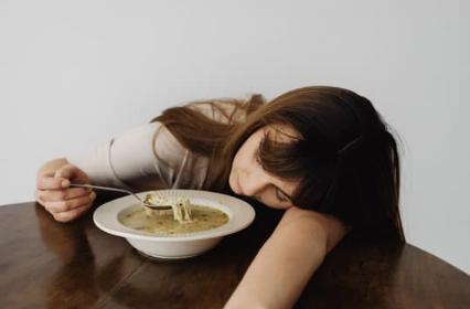 A woman with dark hair resting her head on a table next to a bowl of soup, appearing exhausted or unwell.