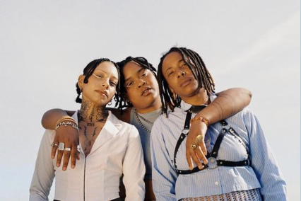 Three women with dreadlocks posing together outdoors against a clear sky, wearing stylish clothing and jewelry.