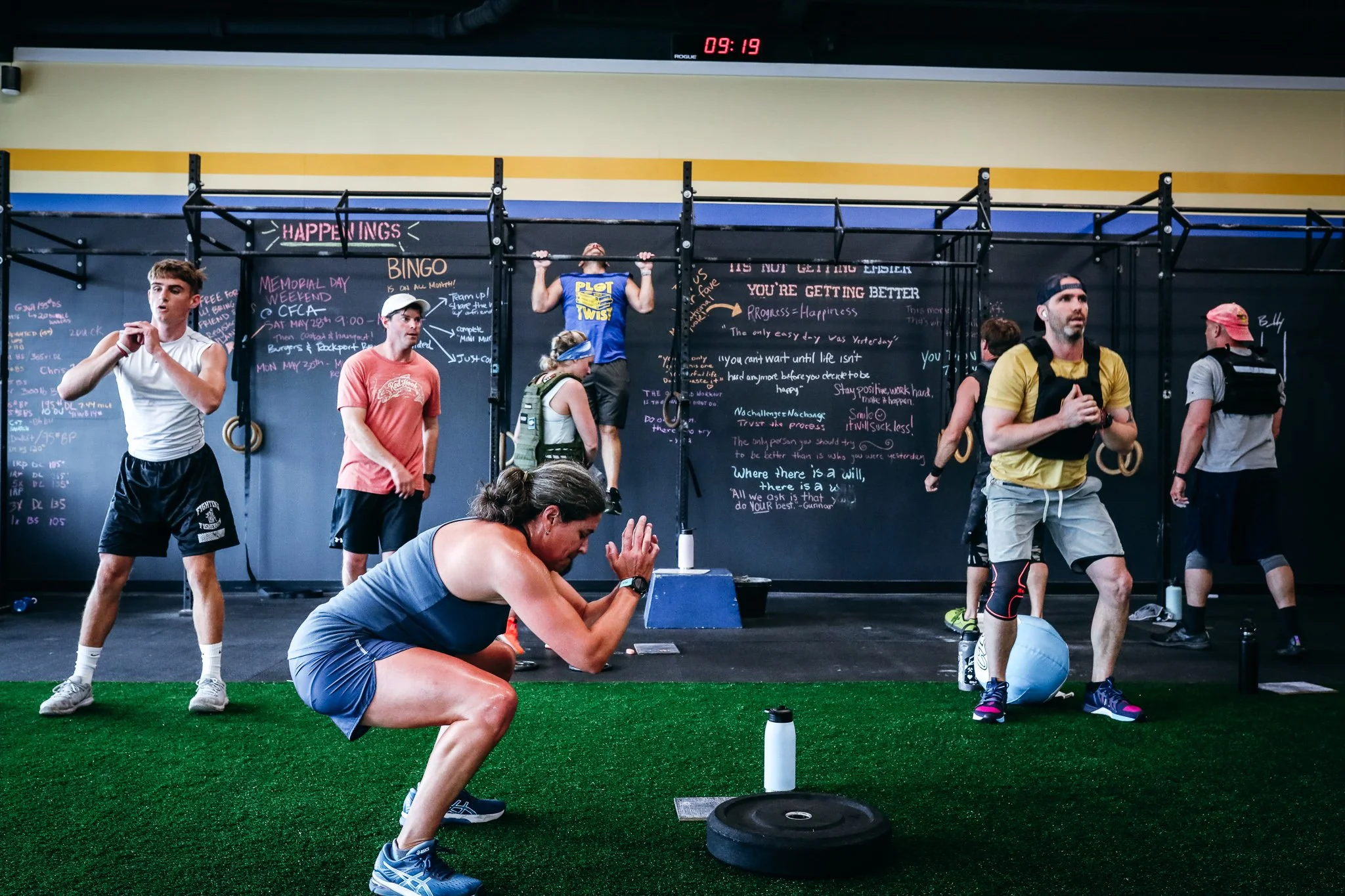 People working out in a gym with chalkboard walls displaying messages and workout schedules, some lifting weights and others preparing for exercises.