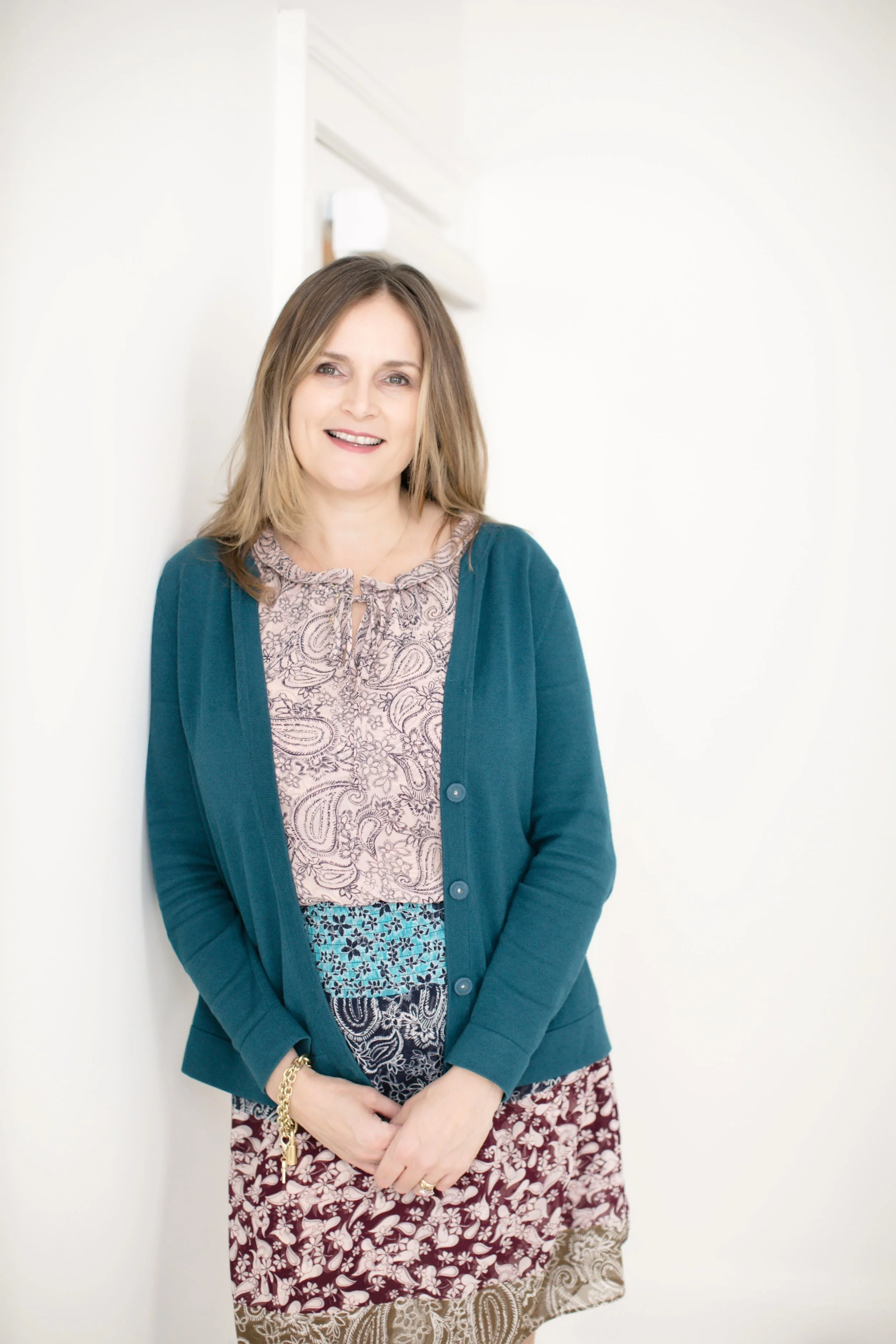Carrie Lettress-Jones wearing a patterned dress and blue cardigan, standing by a white wall background.