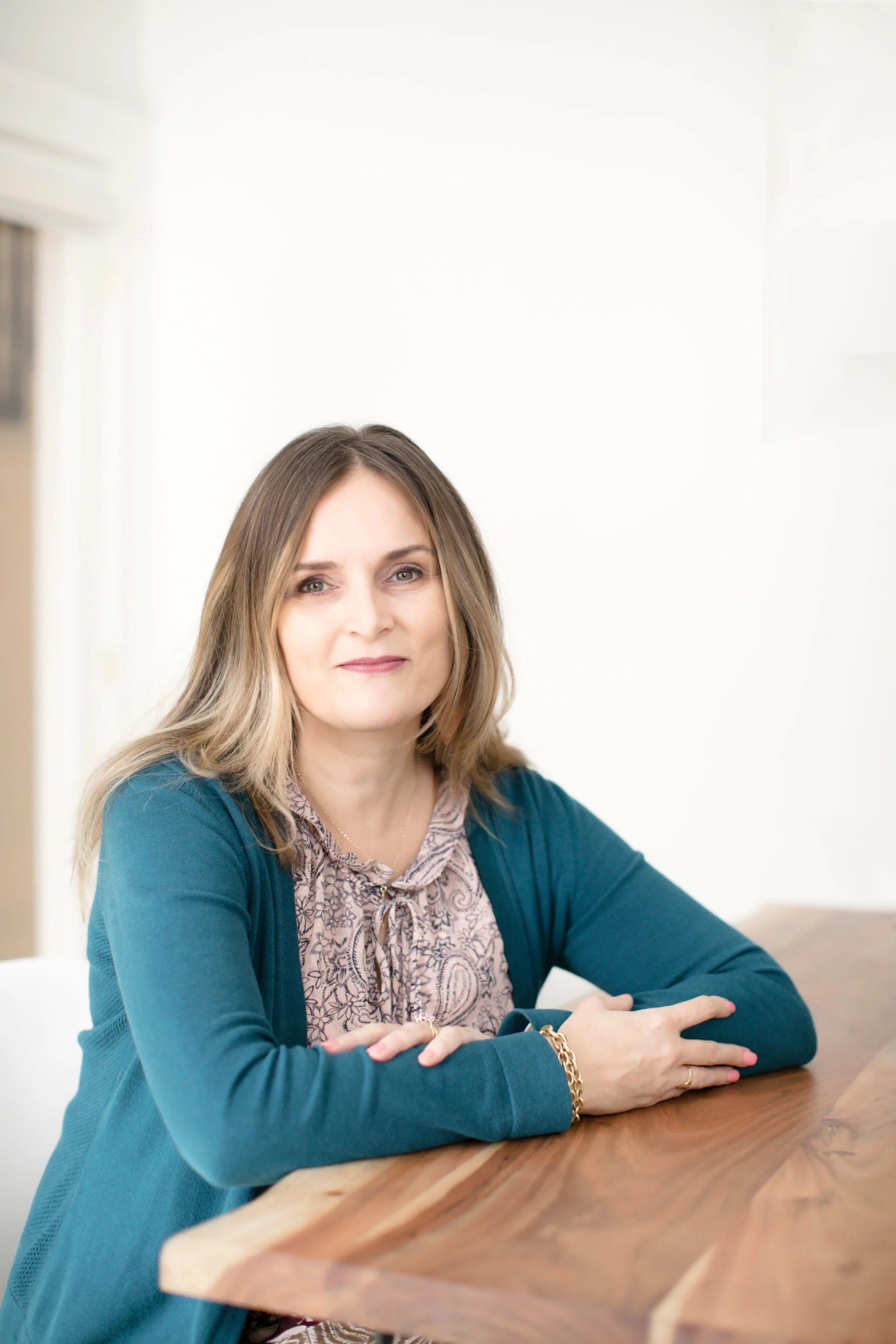 Carrie Lettress-Jones wearing a patterned dress and blue cardigan, sitting at a wooden table in a room with a white wall background.