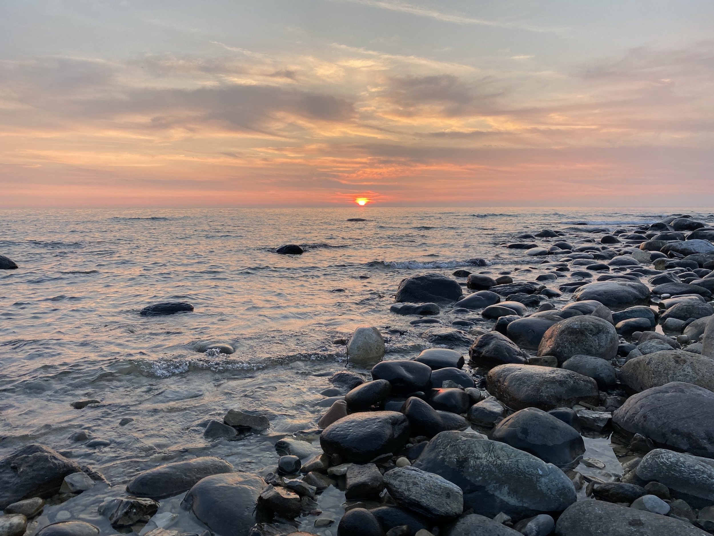 Sunset over the ocean with a colorful sky and clouds, rocky shoreline in the foreground.