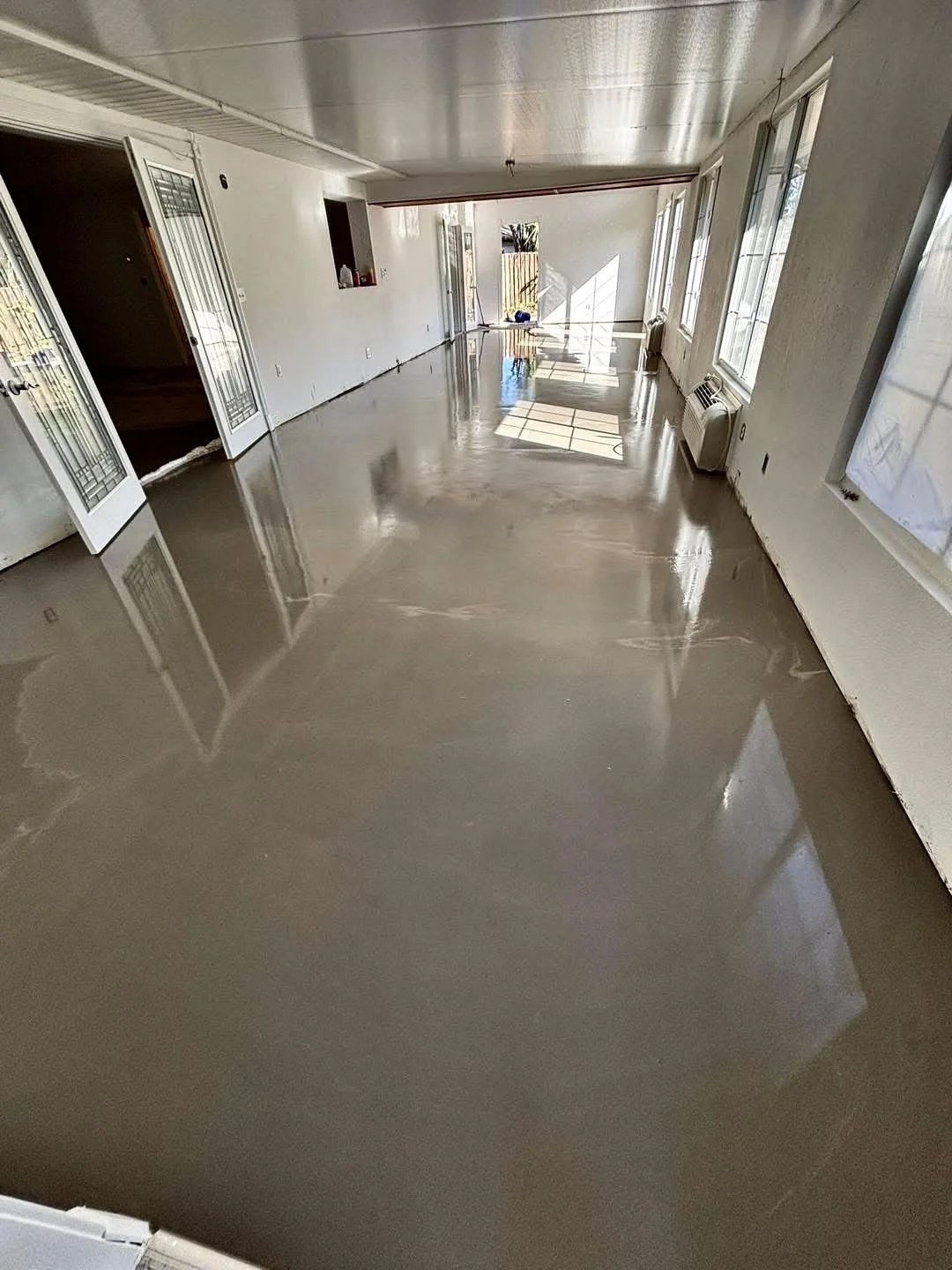 Empty room with freshly polished concrete floors, multiple windows letting in natural light, white walls, and sliding glass door at the far end.