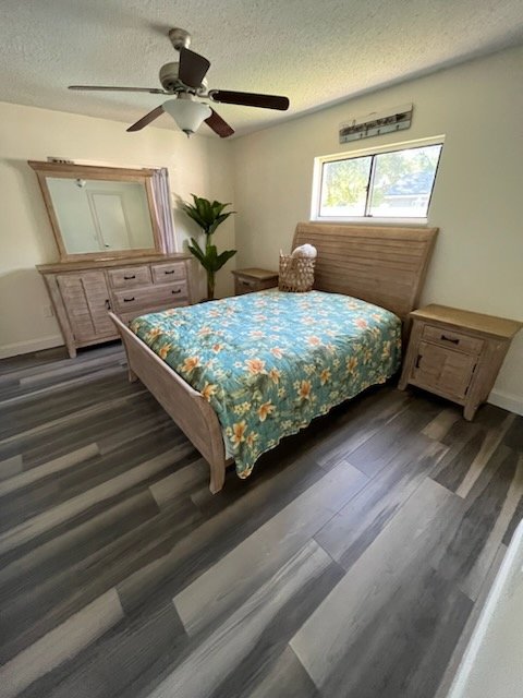 A bedroom with a wooden bed frame and sunflower-patterned bedspread, two matching nightstands, a large mirror on a dresser, a ceiling fan, a window, and a potted plant.
