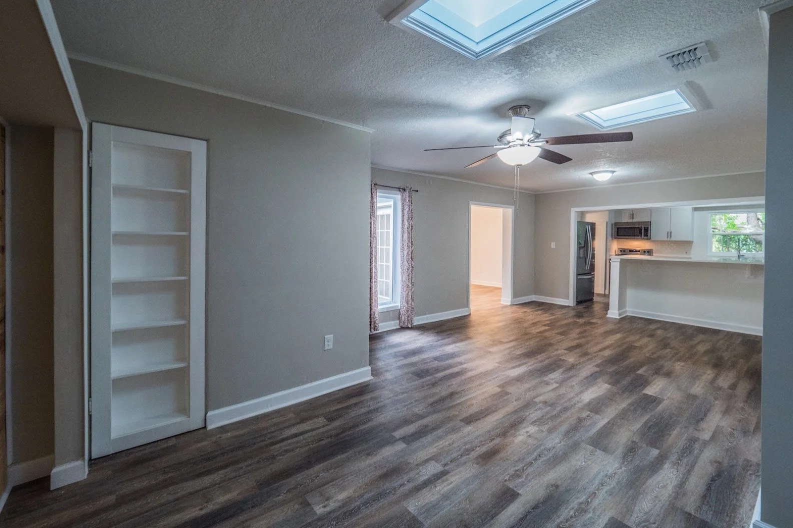 Empty living room with hardwood floors, beige walls, and a ceiling fan. Kitchen area with stainless steel appliances, white cabinets, and a breakfast bar is visible in the background. Skylights and windows let in natural light.